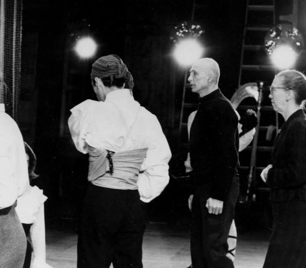 A black and white (archival) photograph from a dance rehearsal, capturing a moment of focused attention among the participants. The bright stage lights in the background create a classic rehearsal ambiance.